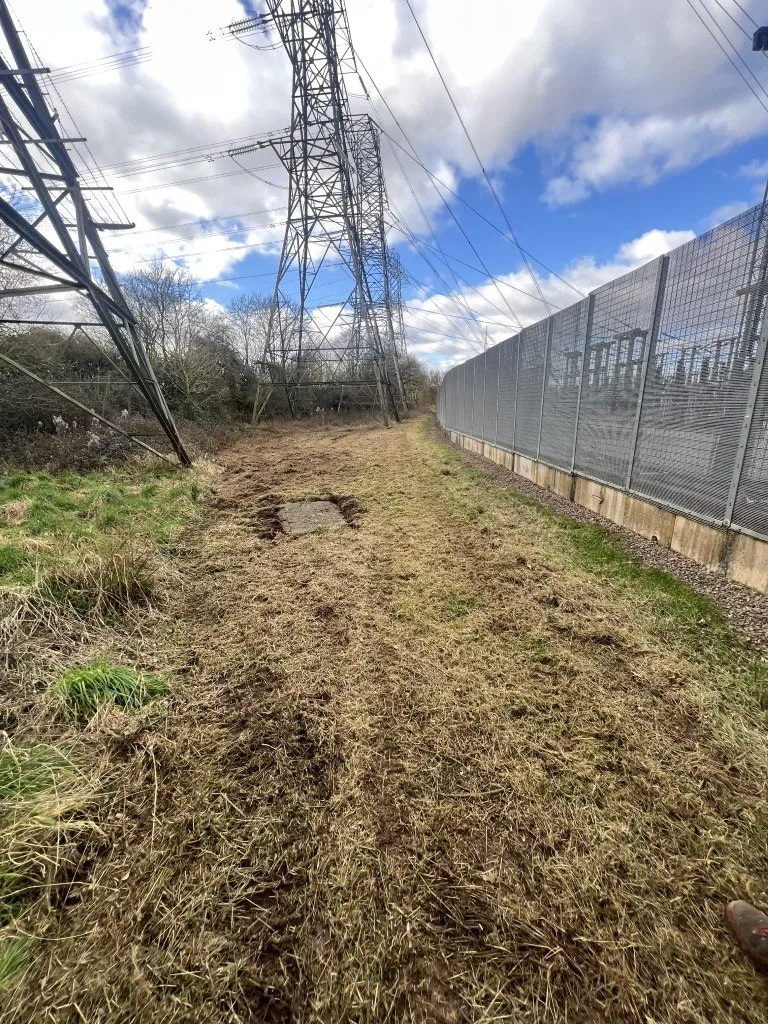 The cable-duct route running alongside the live National Grid substation security fence at Iron Acton, beneath the transmission pylons
